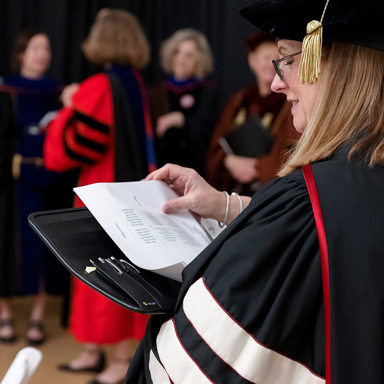 A University Events team member consults a printed sheet in an open leather binder as faculty in formal academic attire gather before an event.